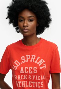 Czerwona bawełniana koszulka z okrągłym dekoltem, krótkimi rękawami i białym napisem "SD SPRINT ACES TRACK & FIELD ATHLETICS" wyraźnie widocznym na przodzie.