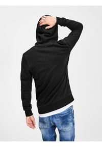 Sort hættetrøje i bomuldsblanding, med ribbede manchetter, afslappet pasform, båret med lysblå denimjeans, synlig hånd med ringe, ensartet baggrund.