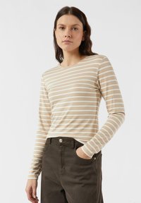 Beżowa bluzka z długim rękawem z białymi poziomymi paskami. Miękka tkanina, okrągły dekolt, dopasowany krój. Połączona z ciemnozielonymi, wysoko taliowanymi spodniami.