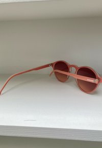 Occhiali da sole rotondi con montatura rosa opaca e lenti sfumate di un rosa scuro. Caratteristiche includono aste solide e dettagli con logo di lato.