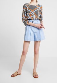 Sort og blå mønstret langærmet top, parret med lyseblå plisserede shorts og lysebrune slidesandaler. Enkel design, glatte teksturer.