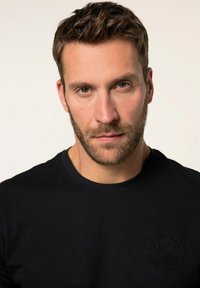 Černé tričko s kulatým výstřihem a texturovaným logem na hrudi, vyrobené z hladkého materiálu a ve fitted designu. Výraz modelu je neutrální.