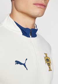 Valge lukuga jope tekstureeritud viimistlusega, millel on tumesinine krae, tumesinine Puma logo ja kuldne embleem rinnal.