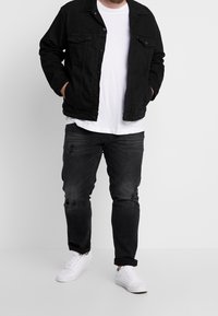 Chaqueta de mezclilla negra con dos bolsillos en la parte delantera, sobre una camiseta blanca. Unos vaqueros negros desgastados y zapatillas blancas completan el conjunto.