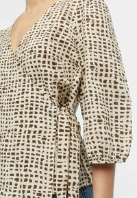 Blouse beige avec un col en V, motif abstrait marron et manches bouffantes. Présente un lien à la taille pour un ajustement réglable.