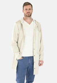 Impermeable de color beige claro con capucha, botones a presión, bolsillo frontal, usado sobre un suéter de punto blanco con escote en V y jeans azules.