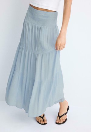 Femme portant une jupe longue à volants bleu clair, un top court blanc et des sandales noires fines, debout devant un fond blanc uni.