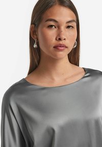 Top grigio seta con scollo ampio e texture liscia, abbinato a orecchini allungati in argento, evidenziando un design contemporaneo.