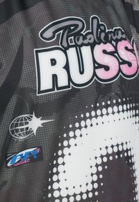 Melnas sporta krekls ar baltu un rozā tekstu "Paolina Russo" un "RUSS." Iezīmes globusa un zvaigznes logo, kā arī punktu raksts dizainā.