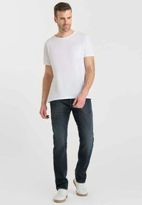 T-shirt blanche à manches courtes avec un détail de poche texturé, associée à un jean bleu foncé et des baskets blanches. Fond uni.