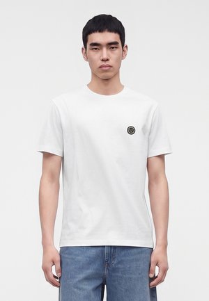 Homme portant un t-shirt blanc uni avec un petit logo noir rond sur la poitrine gauche, associé à un jean bleu, debout devant un fond blanc.