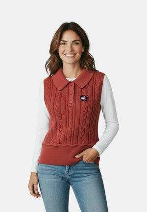Donna sorridente che indossa un gilet senza maniche lavorato a trecce color ruggine con collo sopra una maglia bianca a maniche lunghe e jeans blu.