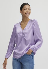 Blouse en satin lavande avec un décolleté en V, détail froncé au centre et poignets élastiqués. Manches longues et coupe décontractée. Associée à un jean.