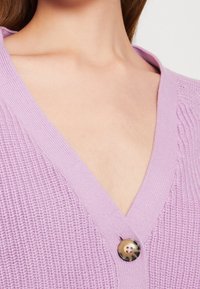 Lila stickad cardigan med djup V-ringning, ribbad textur och en kontrasterande rund knapp med ett marmorliknande mönster.