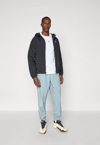 Sort letvægtsjakke med hætte, hvid t-shirt, lyseblå joggers med mesh-detaljer, og sorte og hvide sneakers med orange detaljer.