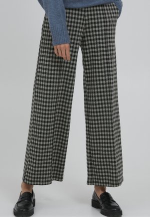 Osoba ubrana w szerokie spodnie w czarno-szarą kratę, niebieski sweter i czarne mokasyny, stojąca na jednolitym tle.