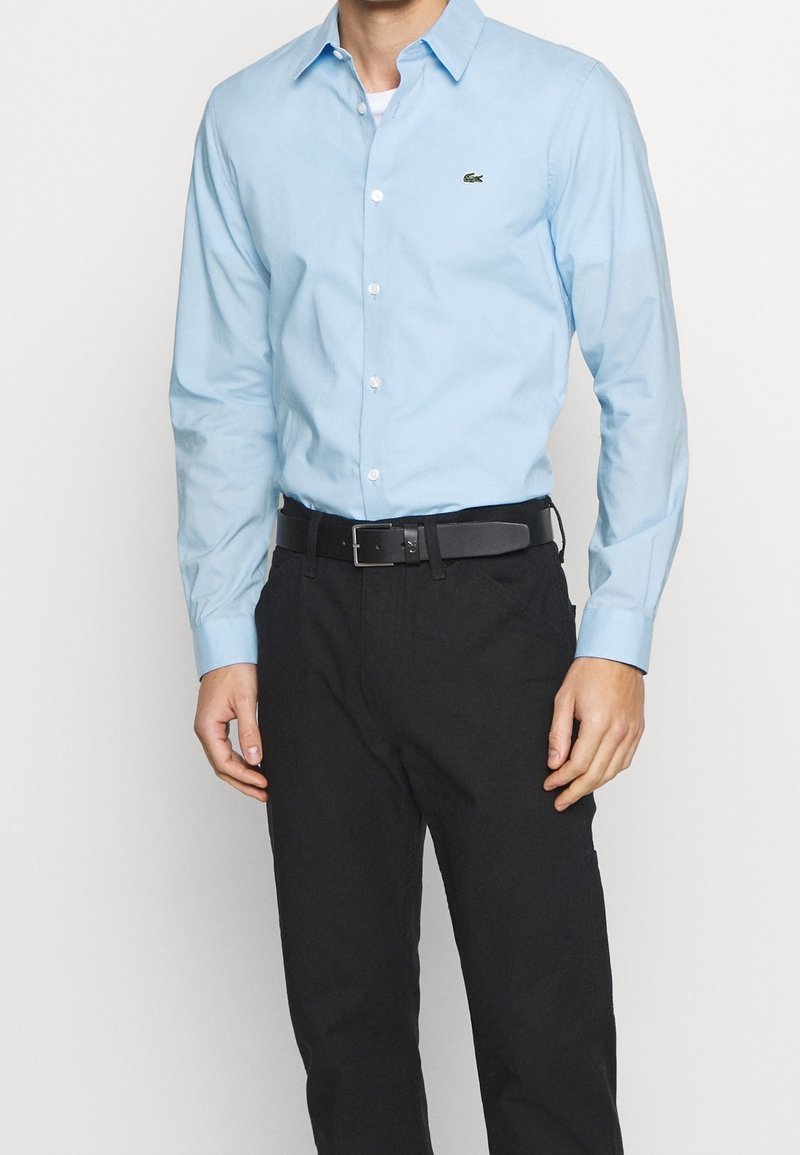 Homme portant une chemise bleu clair boutonnée avec un petit logo sur la poitrine, ceinture noire et pantalon noir, debout devant un fond uni.