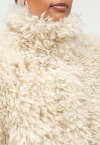 Beżowy, puszysty golf z teksturowanej tkaniny o kręconym wyglądzie; ma wysoki kołnierz i miękką powierzchnię.