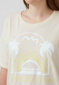 Beige t-shirt met korte mouwen met een witte afbeelding van een zonsopgang achter bergen, met palmbomen en de tekst "GOLDEN HOUR" in het geel.