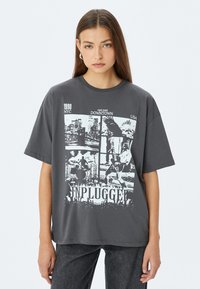 Grijze t-shirt met korte mouwen, voorzien van een grafisch ontwerp van steden en muzikanten, met het woord "UNPLUGGED" prominent weergegeven onderaan.