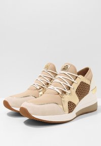 Scarpe da ginnastica beige con accenti in rete testurizzata, dettagli in metallo dorato, lacci bianchi e una suola a cuneo ammortizzata. Materiali in camoscio e sintetico.