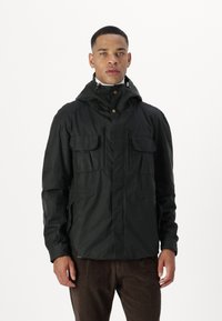 Black, hooded jacket with a zipper and two flap pockets; textured fabric with a slightly rugged look. Worn over brown corduroy pants.