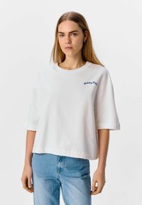 Camiseta blanca de algodón de manga corta con cuello redondo y un diseño corto; presenta texto bordado en azul que dice "Holiday Explore".