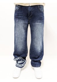 Jeans de mezclilla azul oscuro con un acabado desgastado, corte holgado y un distintivo detalle bordado en la cintura en blanco. Combinados con zapatillas blancas.