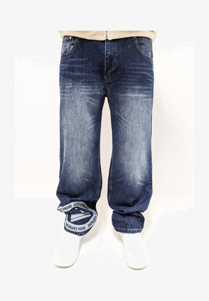 Dunkelblaue Denim-Jeans mit einer ausgewaschenen Optik, lockerer Passform und markantem weißen bestickten Bunddetail. Kombiniert mit weißen Sneakers.