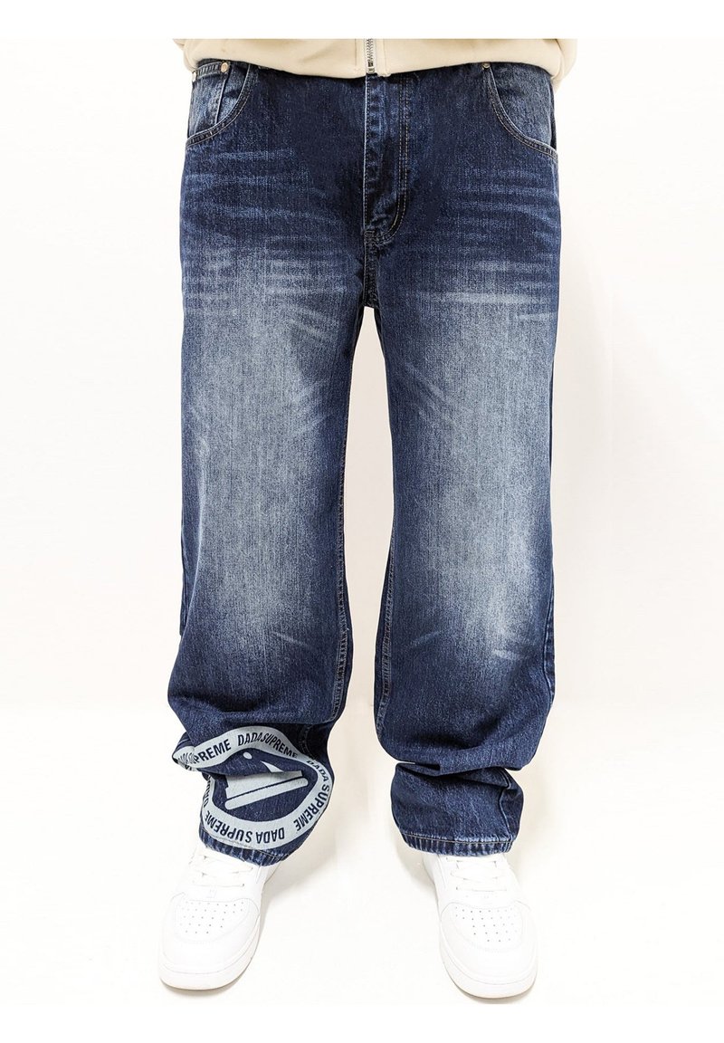 Jeans de mezclilla azul oscuro con un acabado desgastado, corte holgado y un distintivo detalle bordado en la cintura en blanco. Combinados con zapatillas blancas.