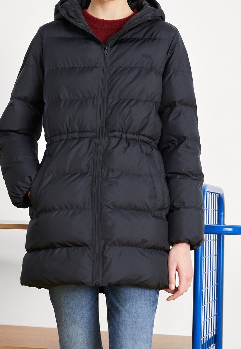 Černá péřová bunda s kapucí, s zipovým zapínáním, stáhnutým pasem a prošívanou texturou. Nosená přes burgundské svetřík a džíny.