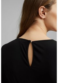 Vêtement noir avec une ouverture en forme de clé à l'arrière, maintenue par un petit détail métallique au-dessus du cou, fabriqué en tissu lisse.