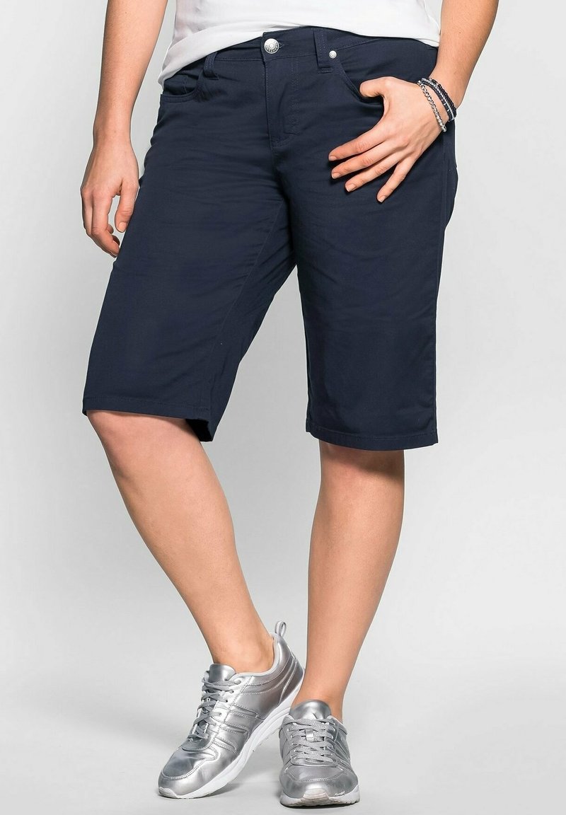 Personne portant un short bleu marine jusqu'aux genoux, une chemise blanche, des baskets argentées et des bracelets au poignet droit sur un fond uni.