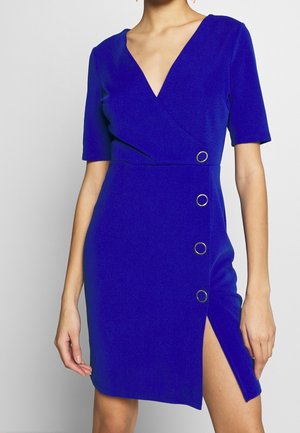 Vrouw draagt een koningsblauwe wikkeljurk met korte mouwen, gouden ronde knopen aan één kant en een hoge split tot op de dij.