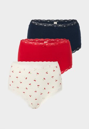 Femme debout pieds nus portant un soutien-gorge blanc et une culotte blanche taille haute à petit motif fleuri rouge, posant une main sur la hanche.