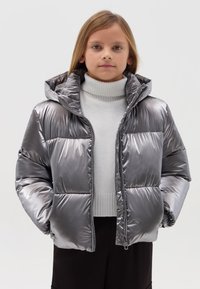 Silberner Puffer-Jacke mit Kapuze, glänzender Oberfläche, dicker Polsterung, Frontreißverschluss und geripptem Kragen, über einem hellen Pullover getragen.