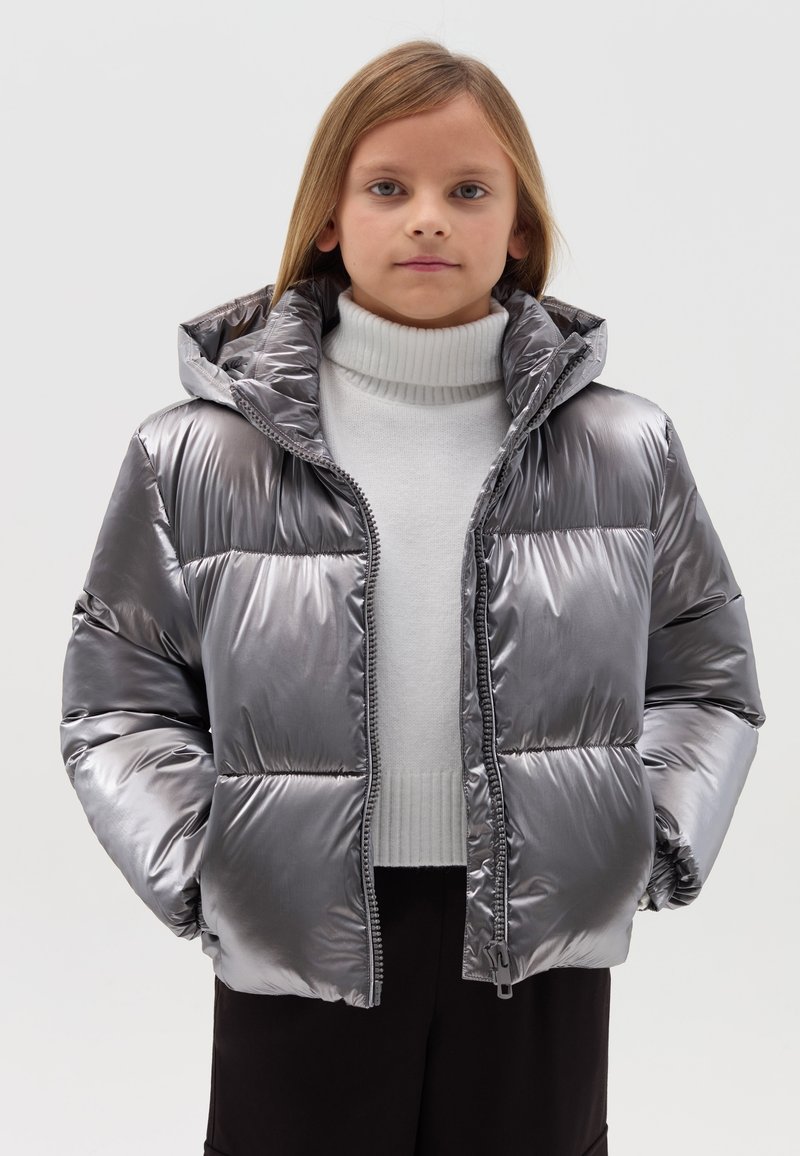 Silberner Puffer-Jacke mit Kapuze, glänzender Oberfläche, dicker Polsterung, Frontreißverschluss und geripptem Kragen, über einem hellen Pullover getragen.