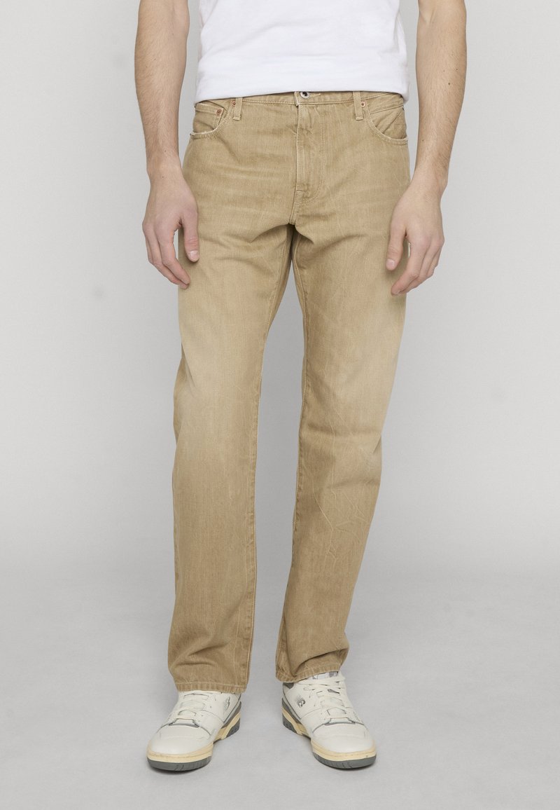 Homme portant un jean beige droit, des baskets blanches et un t-shirt blanc rentré, debout devant un fond clair uni.