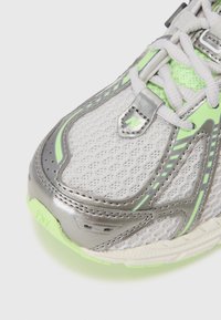 Gros plan d'une chaussure de sport grise et vert clair avec tissu en maille, renforts métalliques et lacets blancs.