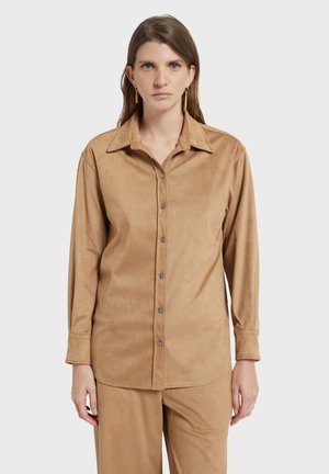 Femme portant une chemise à manches longues en daim couleur beige à boutons, assortie à un pantalon, debout devant un fond gris clair uni.
