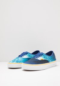 Scarpe da ginnastica in tela blu e turchese con motivi a onda, punta arrotondata, suola bianca e design con lacci blu. Presentano dettagli in giallo e logo.