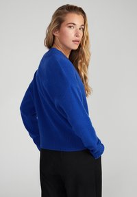 Blauwe gebreide trui met een losse pasvorm, geribbelde manchetten en zoom, en een drop shoulder ontwerp. Draagt over zwarte broek, wat een zachte textuur laat zien.