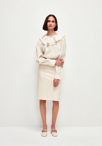 Cremebluse med rynket krave og lange ærmer, parret med en blyantskørt. Modellen bærer lysebeige sko med spænde-detalje.