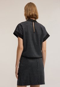 Kobieta z krótkimi jasnobrązowymi włosami, ubrana w czarny wzorzysty top i dopasowaną spódnicę, widziana od tyłu na jednolitym tle.