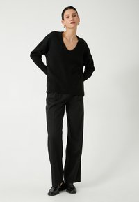 Mustat ribbipaidoilla varustetut syvät V-kaula-aukot, yhdistettynä mustiin leveälahkeisiin housuihin ja mustiin löysiin kenkiin, neutraalia taustaa vasten.