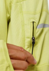 Veste de sport zippée vert citron avec tissu texturé, un accent réfléchissant et un cordon de serrage sur la fermeture éclair pour un accès facile.
