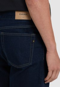 Jeans blu scuro in denim con una texture liscia, dotati di una toppa in pelle beige con l'etichetta "FARAH" e cuciture gialle sulla tasca posteriore.