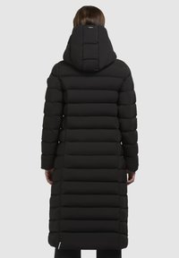 Persoon in een lange zwarte gewatteerde jas met horizontale stiksels en een vaste capuchon, die met de rug naar de camera staat op een effen grijze achtergrond.