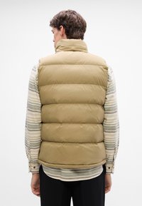 Gilet imbottito beige con colletto alto, motivo trapuntato e finitura liscia, indossato sopra una camicia a maniche lunghe a righe orizzontali.