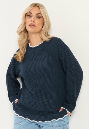 Blonde vrouw die een donkerblauwe gestructureerde trui met witte bies en lichtblauwe spijkerbroek draagt, staand met handen in de zakken tegen een witte achtergrond.